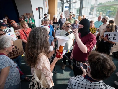 A group of protesters gathers indoors, holding signs and banners, while one person in a black mask speaks into a megaphone held by another protester.