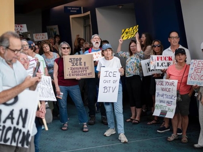 A group of people stand indoors holding protest signs with messages against ICE and supporting refugees and human rights.