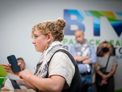 A person with curly hair holds a smartphone while seated at a table; several people stand blurred in the background in front of a wall with a large, colorful "BTV" logo.