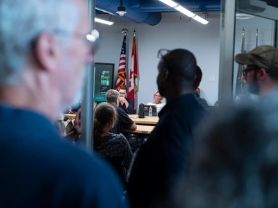 A group of people attends a meeting in a room with American flags; some participants are seated at a table while others observe from the doorway.