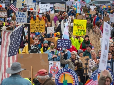 Large crowd at a protest holding various signs related to democracy, leadership, and anti-hate messages; American flags visible among participants.
