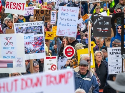 A large crowd of protesters holds signs with messages opposing Trump and promoting resistance, anti-racism, and respect for rule of law at an outdoor demonstration.