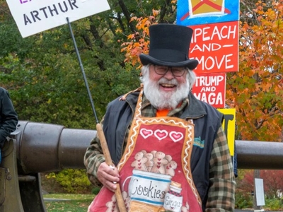A man in a top hat and gingerbread-themed apron holds a sign reading "NO KINGS EXCEPT ARTHUR" at a protest with colorful political signs in the background.