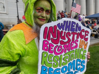 A person in a green costume holds a colorful protest sign reading "When injustice becomes law resistance becomes duty" at a public demonstration outside a large building.