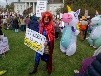 A person in a red cape holds a sign reading "Democracy is our superpower" at an outdoor rally, surrounded by people, some in inflatable unicorn costumes and holding protest signs.