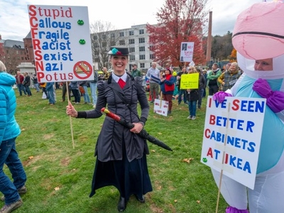 A person in a Mary Poppins costume holds a protest sign among a crowd; another person in a unicorn costume holds a sign referencing cabinets from Ikea.