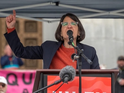 A woman speaks passionately into a microphone at a podium during an outdoor event, raising her left hand with her index finger pointed upward.