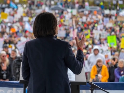A person speaks at a podium to a large crowd holding signs at an outdoor event or protest.