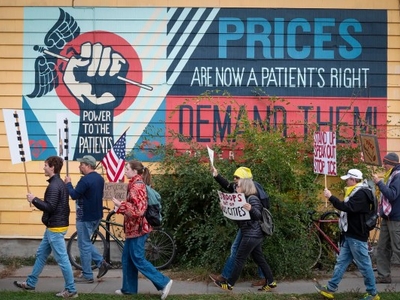 A group of people holding protest signs walks past a mural that reads, “PRICES ARE NOW A PATIENT’S RIGHT. POWER TO THE PATIENTS. DEMAND THEM!”.