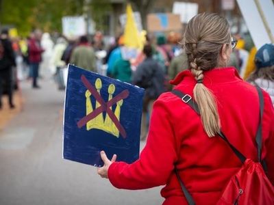 A person in a red coat holds a sign with a yellow crown crossed out at a protest or demonstration, with other participants and signs visible in the background.