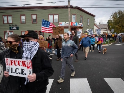A group of protesters march on a street, holding signs and an American flag; one prominent sign reads "FUCK I.C.E." while others display various messages.