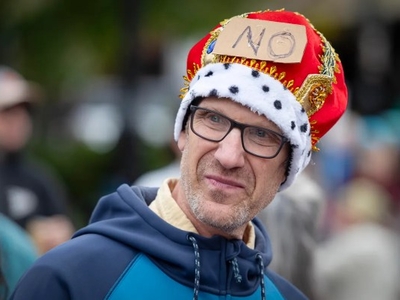 A man wearing glasses and a red crown with a cardboard sign reading "NO" attached to it stands outdoors, looking to the side.