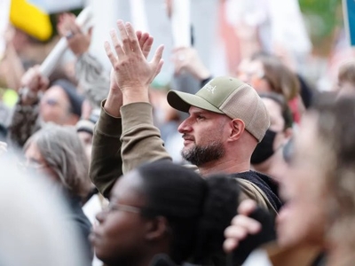 A man in a green cap claps while standing among a crowd of people at a public gathering or protest.