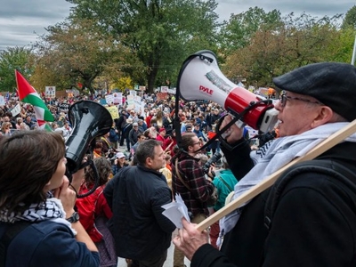 A large crowd gathers outdoors for a protest; people hold signs and speak through megaphones, with flags and trees visible in the background.