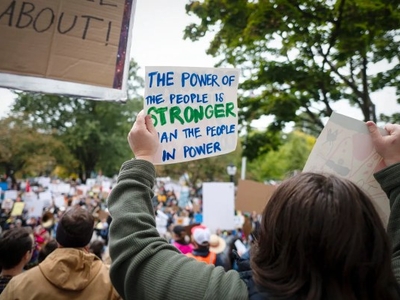A person holds a sign reading "The power of the people is stronger than the people in power" at a crowded outdoor protest.
