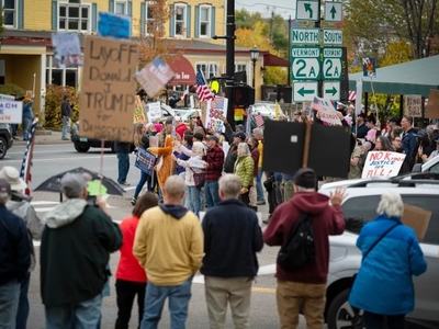 A crowd of people gathers at an intersection holding protest signs, with buildings, street signs, and trees visible in the background.