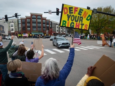 A crowd of people protest at a city intersection, holding signs and American flags. One prominent sign reads "Defend Your Rights" in large, colorful letters.