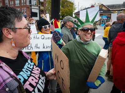 A group of people protest on a city street, one dressed as the Statue of Liberty holding a sign, others holding protest signs in support of trans kids.