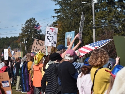 A group of people stand outdoors holding protest signs and banners, with trees and power lines visible in the background.