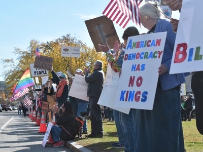 A group of people stand and kneel along a sidewalk holding protest signs and American flags; one sign reads "AMERICAN DEMOCRACY HAS NO KINGS.