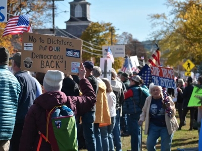 A group of people protest outdoors, holding signs with messages including “No to Dictators, Put Our Democracy Back!” and “Stop Trump Save Democracy.