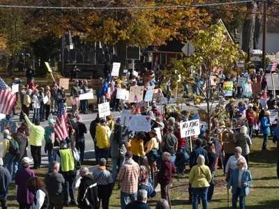 A large group of people gathers outdoors holding various protest signs and American flags near a street lined with trees in autumn.