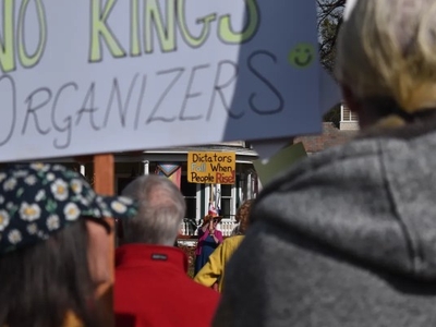People at an outdoor protest hold signs, including one in the background that reads “Dictators Fall When People Rise.”.
