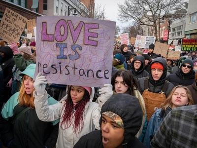 A crowd of people at a protest march, with one person in the foreground holding a sign that reads “LOVE IS resistance.” Other signs and winter clothing are visible.