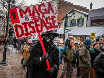 A person in dark clothing and a mask holds a red sign reading "MAGA IS A DISEASE" during a crowded street protest; other protesters stand and walk behind them.
