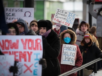 A group of protesters hold signs opposing ICE, with messages like “Refuse ICE,” “ICE out now,” and “America has no king.” One person wears a mask.