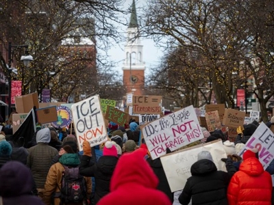 A crowd of people holding protest signs march down a decorated street lined with trees and buildings, with a church visible in the background.
