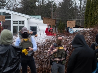 Protesters hold "No ICE" signs outside a house as law enforcement officers wearing "POLICE ICE" and "ERO" vests stand in the foreground.