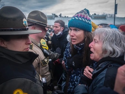 Two police officers face a crowd of people outdoors; one older woman is crying while another woman speaks to an officer.