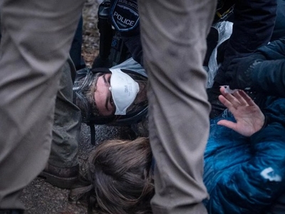 A person wearing a face mask lies on the ground looking up, surrounded by police officers; another person’s hand and head are visible nearby.