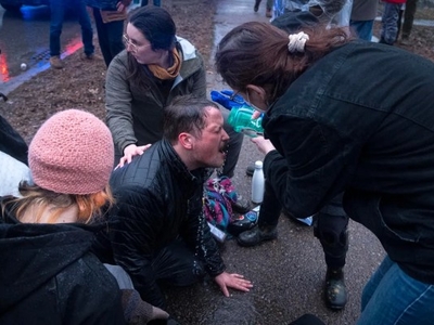 A person kneels on the ground with eyes closed while another pours liquid on their face; others surround and support them outdoors.