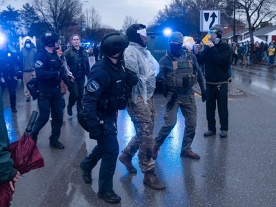 Police officers in tactical gear detain and escort a person in plastic restraints during a street protest, with onlookers and emergency lights visible in the background.
