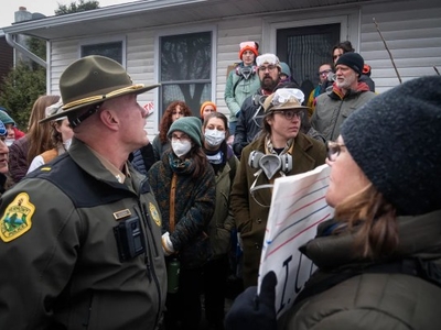 A group of people, some wearing masks and holding signs, stand outside a house while speaking with uniformed police officers.