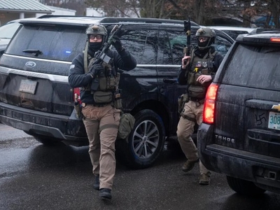 Two armed law enforcement officers in tactical gear walk between SUVs on a wet street, appearing to respond to an active situation.