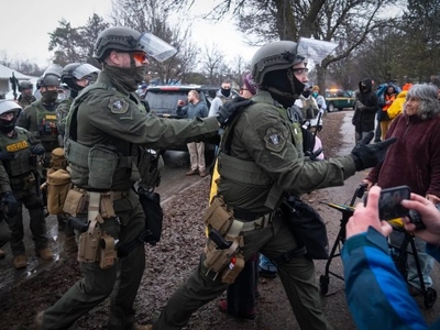 Police officers in tactical gear move through a crowd on a muddy street, with bystanders and vehicles in the background on a cloudy day.