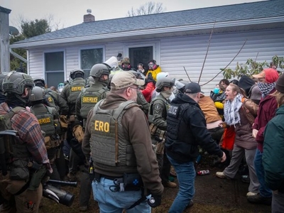 Law enforcement officers in tactical gear confront a group of protesters outside a white house, with some people shouting and others observing.