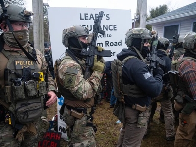 A group of heavily armed police officers in tactical gear stand in formation outdoors near a sign and a crowd.