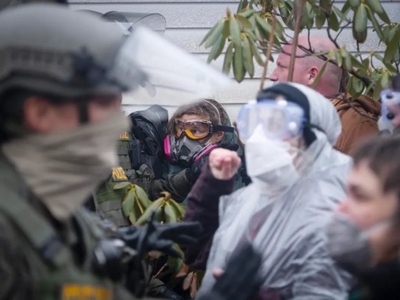 Law enforcement officers in riot gear face protesters wearing masks and goggles. Some protesters wear plastic coverings and appear to be shouting or gesturing.