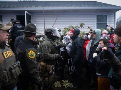 Police officers in tactical gear face a crowd of protesters outside a house; some protesters hold signs and take photos while officers block the entrance.
