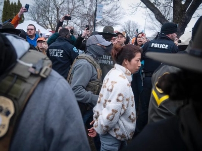 A woman in a white patterned jacket is handcuffed and escorted by law enforcement officers through a crowd of onlookers, some of whom are recording with their phones.