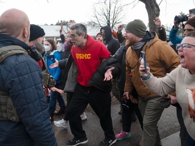 A group of people confronts law enforcement officers outdoors; some individuals appear to be shouting or recording on their phones.