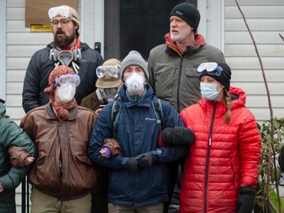 A group of people in winter clothing and protective masks stand arm-in-arm outside a building, wearing goggles and face coverings.