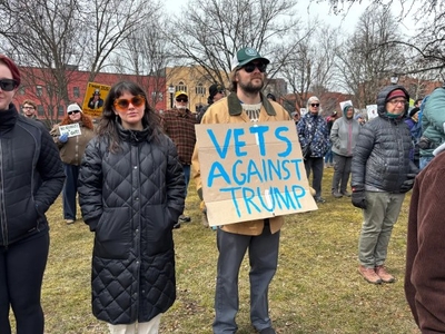 A group of people stand outdoors at a protest. One person holds a cardboard sign reading "Vets Against Trump." Trees without leaves and buildings are visible in the background.