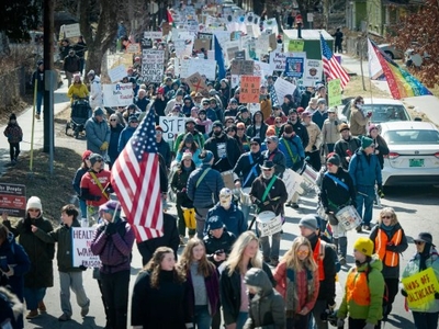 A large group of people walks down a street holding signs and flags during a protest or march on a sunny day.