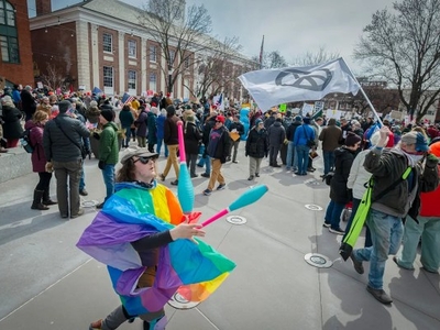 A person in a rainbow flag juggles clubs in front of a large crowd gathered outside, with some people holding signs and flags.