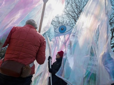 People hold translucent banners and a large sign with a blue eye design during an outdoor gathering on a sunny day, with bare trees in the background.